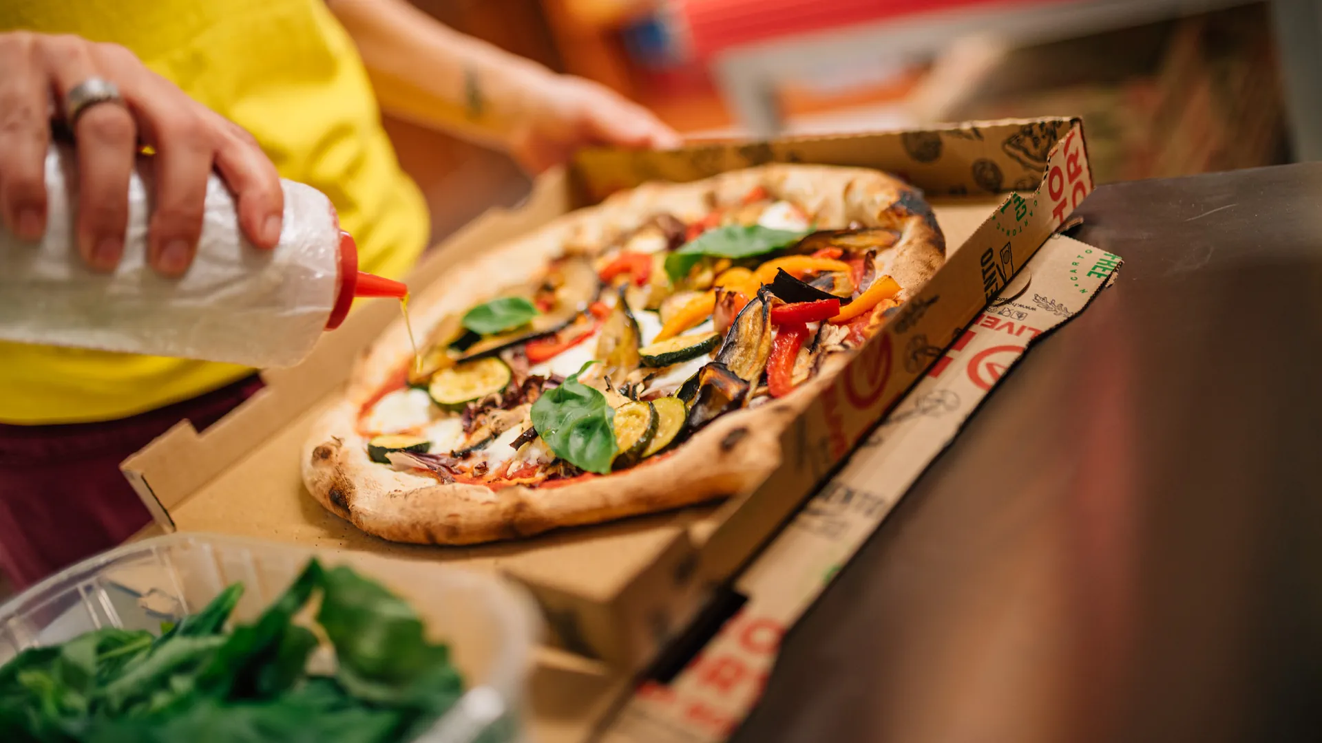 Una pizza con verdure grigliate nel cartone, mentre si aggiunge l'olio prima della consegna