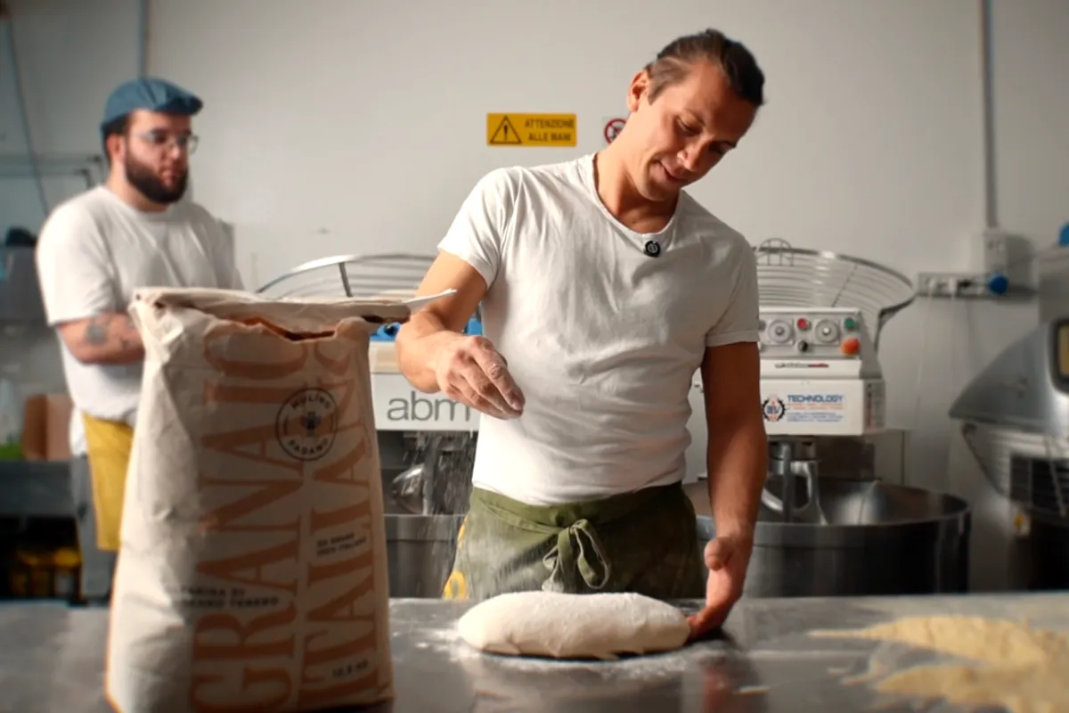 Il pizzaiolo lavora un panetto di impasto sul banco d'acciaio del laboratorio, accanto al sacco di farina